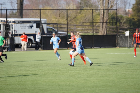 A group of female soccer players are actively engaged in a match on a green field. Two players in blue uniforms are challenging a player in an orange and blue uniform for the ball. Officials and support staff can be seen in the background, one wearing a red shirt. An ambulance is parked near the fence.