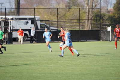 A group of female soccer players are actively engaged in a match on a green field. Two players in blue uniforms are challenging a player in an orange and blue uniform for the ball. Officials and support staff can be seen in the background, one wearing a red shirt. An ambulance is parked near the fence.