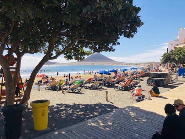 A bustling beach scene with numerous people lounging on sunbeds under bright umbrellas. The beach is bordered by a wooden boardwalk and shaded by leafy trees. In the background, a mountain rises near the ocean, and a clear, sunny sky dominates the scene.