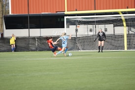 Two soccer players, one in a blue jersey and the other in a red and blue striped jersey, are actively engaging in a match near the goalpost. The player in red is attempting a sliding tackle, while the player in blue is focusing on controlling the ball. A goalkeeper in black stands ready in front of the net, and another player observes from the sidelines.
