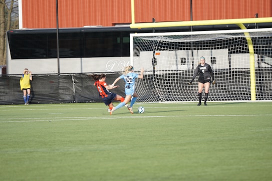 Two soccer players, one in a blue jersey and the other in a red and blue striped jersey, are actively engaging in a match near the goalpost. The player in red is attempting a sliding tackle, while the player in blue is focusing on controlling the ball. A goalkeeper in black stands ready in front of the net, and another player observes from the sidelines.