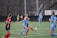 Players engaged in a competitive soccer match on a green field. One player in a blue uniform is actively kicking the ball while another player in a red and black uniform attempts to block. Two additional players in blue uniforms and another player in a red uniform are closely observing the action. The background shows others standing near the sideline.
