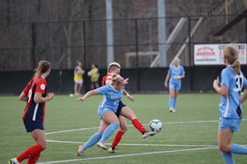 Players engaged in a competitive soccer match on a green field. One player in a blue uniform is actively kicking the ball while another player in a red and black uniform attempts to block. Two additional players in blue uniforms and another player in a red uniform are closely observing the action. The background shows others standing near the sideline.