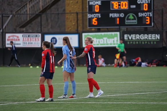 Soccer players stand on a field, with two wearing red and blue uniforms and one in a light blue uniform. A scoreboard in the background shows a score of zero for both teams. A referee in green is visible near the sidelines along with other players resting and some advertisements visible on the fencing.