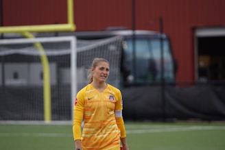 A person wearing a yellow soccer jersey stands on a sports field with a goal post and net in the background. The individual has long hair tied back and appears to be casually walking or standing on the field. A large bus and a red building are visible in the background.