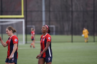 Two women in red and navy soccer jerseys are walking on a soccer field. In the background, another player is blurred, wearing a similar uniform. A soccer goalpost and net are visible in the distance, with a goalkeeper dressed in yellow standing near the goal.