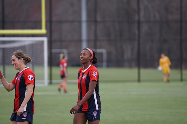 Two women in red and navy soccer jerseys are walking on a soccer field. In the background, another player is blurred, wearing a similar uniform. A soccer goalpost and net are visible in the distance, with a goalkeeper dressed in yellow standing near the goal.
