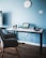 A modern workspace features a sleek, minimalist desk against a light blue wall. On the desk, a laptop with a screen displaying architectural designs sits open. Surrounding the laptop are two camera lenses, a mug, a small camera, and headphones. A clock with a simple design is mounted on the wall, complementing the room's clean, organized aesthetic. The wooden floor adds a warm touch to the overall setting.
