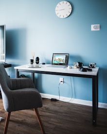 A modern workspace features a sleek, minimalist desk against a light blue wall. On the desk, a laptop with a screen displaying architectural designs sits open. Surrounding the laptop are two camera lenses, a mug, a small camera, and headphones. A clock with a simple design is mounted on the wall, complementing the room's clean, organized aesthetic. The wooden floor adds a warm touch to the overall setting.
