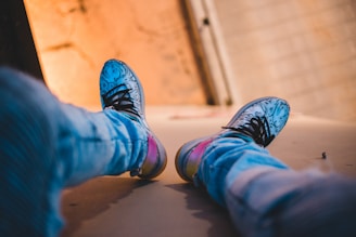 A pair of brightly colored sneakers with artistic patterns is worn by someone sitting with legs extended. The person is wearing light blue jeans. The angle suggests the person is sitting on an elevated surface with a blurred background showing a warm, orange and beige wall.
