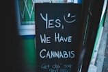 A chalkboard sign with the message 'Yes, we have cannabis' and a smiley face drawn above it. It is propped up outside a building with green walls.