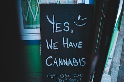 A chalkboard sign with the message 'Yes, we have cannabis' and a smiley face drawn above it. It is propped up outside a building with green walls.