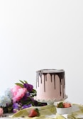 A pink frosted cake with chocolate drip icing sits on a white cake stand. Surrounding the cake are fresh flowers in shades of purple and pink, lying on a green and white checkered cloth. There are also a few strawberries, one whole and one sliced, placed near the cake, with the sliced one in a white bowl.