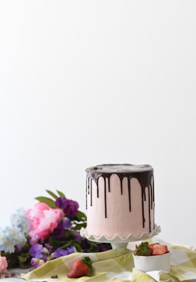 A pink frosted cake with chocolate drip icing sits on a white cake stand. Surrounding the cake are fresh flowers in shades of purple and pink, lying on a green and white checkered cloth. There are also a few strawberries, one whole and one sliced, placed near the cake, with the sliced one in a white bowl.