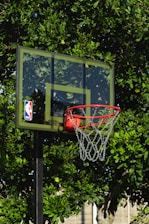 A basketball hoop with a transparent backboard adorned with an NBA logo stands in front of lush green foliage. The vibrant leaves create a natural backdrop for the outdoor basketball setup.