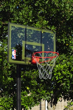 A basketball hoop with a transparent backboard adorned with an NBA logo stands in front of lush green foliage. The vibrant leaves create a natural backdrop for the outdoor basketball setup.