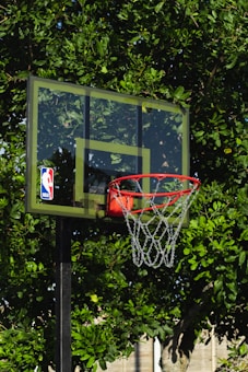 A basketball hoop with a transparent backboard adorned with an NBA logo stands in front of lush green foliage. The vibrant leaves create a natural backdrop for the outdoor basketball setup.