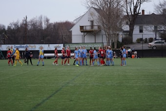 A group of female soccer players is standing on a green field. Two teams wearing different uniforms, one in red and black, and the other in blue, appear to be interacting before or after a game. There is a row of advertising boards and a residential area with trees in the background.