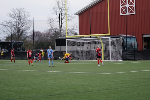 A group of soccer players on a field with a goalpost in the background. The players are actively engaged in a game, wearing distinctive uniforms with one team in red and black and the other in blue. The setting appears to be an outdoor soccer field with buses and a barn-like structure visible in the background.
