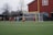 A group of soccer players on a field with a goalpost in the background. The players are actively engaged in a game, wearing distinctive uniforms with one team in red and black and the other in blue. The setting appears to be an outdoor soccer field with buses and a barn-like structure visible in the background.