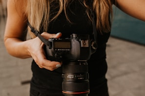 A person holding a DSLR camera with both hands, wearing a black sleeveless top. The camera appears to be a professional model with a large lens, and a camera strap is attached.