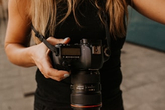 A person holding a DSLR camera with both hands, wearing a black sleeveless top. The camera appears to be a professional model with a large lens, and a camera strap is attached.