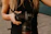 A person holding a DSLR camera with both hands, wearing a black sleeveless top. The camera appears to be a professional model with a large lens, and a camera strap is attached.