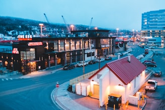 A busy urban setting featuring restaurants with illuminated signs, surrounded by parked cars. The foreground has a white building with a red roof, while the background reveals a cityscape with lit structures and a few cranes in the distance.