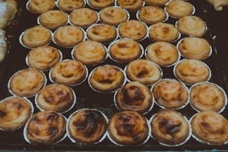 A display of numerous small pastries arranged in neat rows, each in its own metallic baking cup. The pastries have a golden-brown, flaky crust with some baked areas appearing slightly darker.