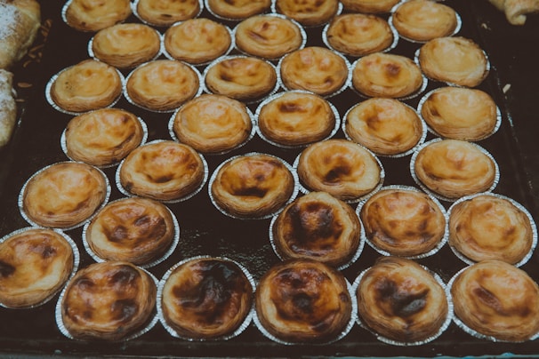 A display of numerous small pastries arranged in neat rows, each in its own metallic baking cup. The pastries have a golden-brown, flaky crust with some baked areas appearing slightly darker.