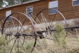 An old, rusted agricultural machinery with large metal wheels is situated in front of a wooden building. Tall, dry grass surrounds the machinery, adding to the rustic and abandoned atmosphere. The wooden building behind has several windows and blends with the rural setting.