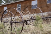 An old, rusted agricultural machinery with large metal wheels is situated in front of a wooden building. Tall, dry grass surrounds the machinery, adding to the rustic and abandoned atmosphere. The wooden building behind has several windows and blends with the rural setting.