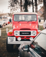 A bright red vintage Toyota Land Cruiser is parked outdoors, with several other cars lined up behind it. The setting appears to be a park or outdoor event area with grass and tall trees in the background. The vehicle's distinct front design and vibrant color make it stand out as the central focus of the image.
