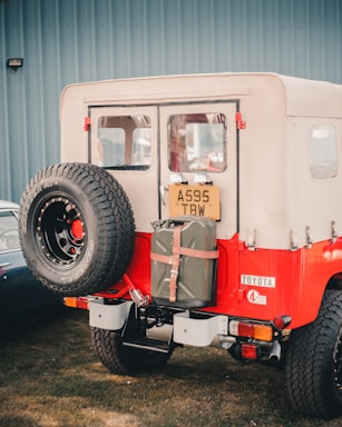 A vintage red and white off-road vehicle with a spare tire mounted on the rear door. A green fuel can attached to the back and a car parked next to it. The ground is covered with grass and a blue building wall is in the background.