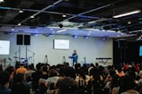A speaker stands on a stage in front of an audience seated in rows. Musical instruments, such as a drum set and keyboards, are visible on the sides. The room is equipped with stage lighting and large screens that display text. The atmosphere suggests a seminar or presentation setting.