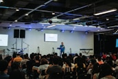 A speaker stands on a stage in front of an audience seated in rows. Musical instruments, such as a drum set and keyboards, are visible on the sides. The room is equipped with stage lighting and large screens that display text. The atmosphere suggests a seminar or presentation setting.