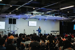 A speaker stands on a stage in front of an audience seated in rows. Musical instruments, such as a drum set and keyboards, are visible on the sides. The room is equipped with stage lighting and large screens that display text. The atmosphere suggests a seminar or presentation setting.