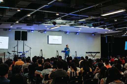 A speaker stands on a stage in front of an audience seated in rows. Musical instruments, such as a drum set and keyboards, are visible on the sides. The room is equipped with stage lighting and large screens that display text. The atmosphere suggests a seminar or presentation setting.