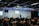 A speaker stands on a stage in front of an audience seated in rows. Musical instruments, such as a drum set and keyboards, are visible on the sides. The room is equipped with stage lighting and large screens that display text. The atmosphere suggests a seminar or presentation setting.