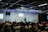A speaker stands on a stage in front of an audience seated in rows. Musical instruments, such as a drum set and keyboards, are visible on the sides. The room is equipped with stage lighting and large screens that display text. The atmosphere suggests a seminar or presentation setting.