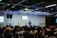 A speaker stands on a stage in front of an audience seated in rows. Musical instruments, such as a drum set and keyboards, are visible on the sides. The room is equipped with stage lighting and large screens that display text. The atmosphere suggests a seminar or presentation setting.