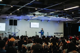 A speaker stands on a stage in front of an audience seated in rows. Musical instruments, such as a drum set and keyboards, are visible on the sides. The room is equipped with stage lighting and large screens that display text. The atmosphere suggests a seminar or presentation setting.