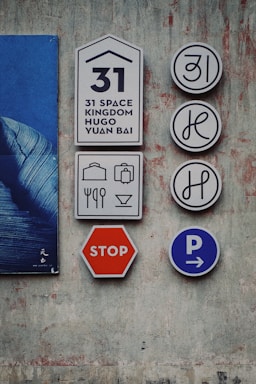 The image depicts several signs mounted on a textured wall. There is a set of signs with the number 31, and graphics representing possibly a hotel or guest service notation. Accompanying them are icons for luggage, utensils, and drinks, suggesting services like accommodation or dining. A red stop sign and a blue parking sign with arrows hint at transportation or traffic indications. The wall is painted with a distressed, artistic finish, and part of a blue poster is visible.
