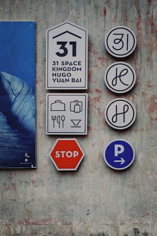 The image depicts several signs mounted on a textured wall. There is a set of signs with the number 31, and graphics representing possibly a hotel or guest service notation. Accompanying them are icons for luggage, utensils, and drinks, suggesting services like accommodation or dining. A red stop sign and a blue parking sign with arrows hint at transportation or traffic indications. The wall is painted with a distressed, artistic finish, and part of a blue poster is visible.