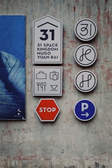 The image depicts several signs mounted on a textured wall. There is a set of signs with the number 31, and graphics representing possibly a hotel or guest service notation. Accompanying them are icons for luggage, utensils, and drinks, suggesting services like accommodation or dining. A red stop sign and a blue parking sign with arrows hint at transportation or traffic indications. The wall is painted with a distressed, artistic finish, and part of a blue poster is visible.