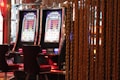 A pair of casino slot machines, each with detailed screens displaying various game outcomes and jackpot amounts. The chairs in front of the slot machines are dark and cushioned for comfort. The background features a hanging curtain made of golden ropes and other casino decorations are visible.