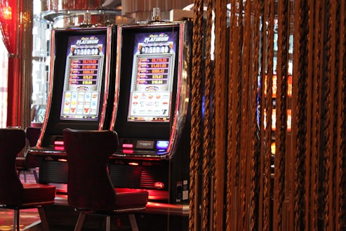 A pair of casino slot machines, each with detailed screens displaying various game outcomes and jackpot amounts. The chairs in front of the slot machines are dark and cushioned for comfort. The background features a hanging curtain made of golden ropes and other casino decorations are visible.