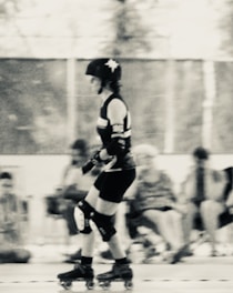 A person is roller skating indoors, wearing protective gear including a helmet, elbow pads, and knee pads. The scene is slightly blurred, giving a sense of movement. The background is out of focus, with several seated individuals and a mesh fence visible.