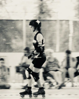 A person is roller skating indoors, wearing protective gear including a helmet, elbow pads, and knee pads. The scene is slightly blurred, giving a sense of movement. The background is out of focus, with several seated individuals and a mesh fence visible.
