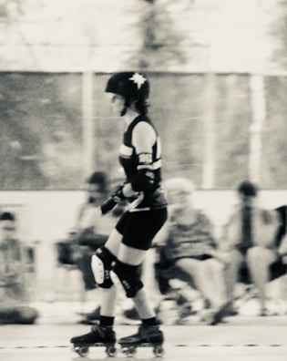 A person is roller skating indoors, wearing protective gear including a helmet, elbow pads, and knee pads. The scene is slightly blurred, giving a sense of movement. The background is out of focus, with several seated individuals and a mesh fence visible.
